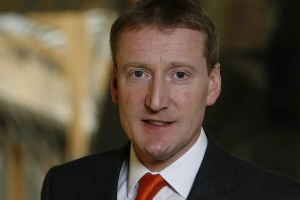 The image shows Tavish Scott facing at the camera and smiling in front of a brown background
