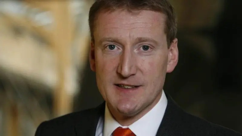 The image shows Tavish Scott facing at the camera and smiling in front of a brown background