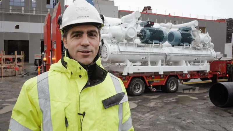 Arild Vikan, project manager at Salmon Evolution, in hi viz and hard hat standing in front of a heat pump being transported on a lorry