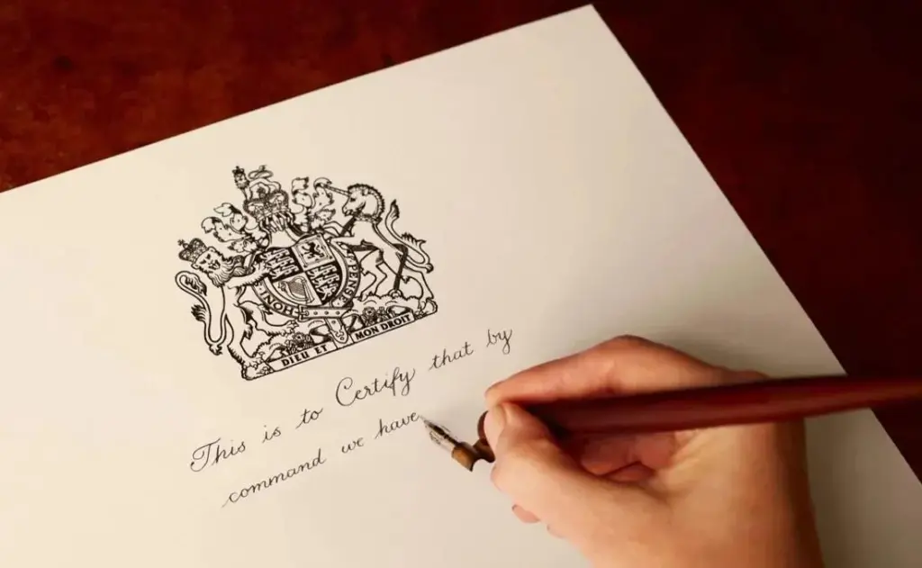 A hand holding a pen and writing on a royal warrant. Text says 'This is to certify that by command we have'