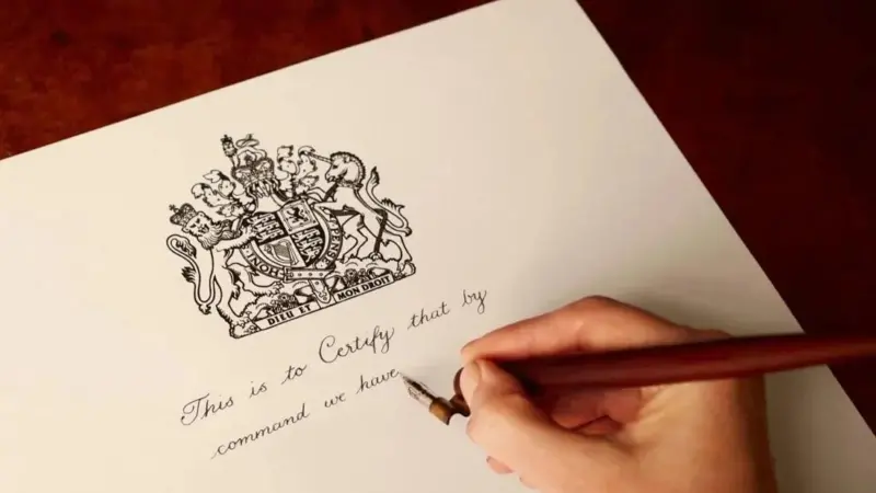 A hand holding a pen and writing on a royal warrant. Text says 'This is to certify that by command we have'