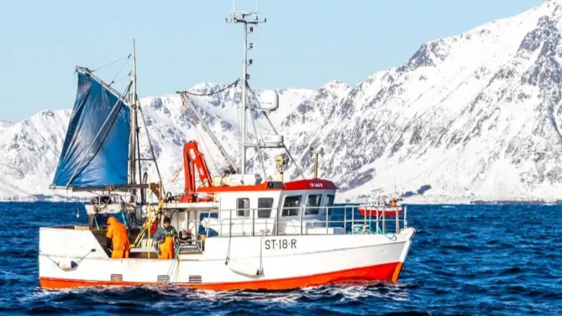 The image shows a Norwegian fishing vessel on the water with the snowy coastline in the background