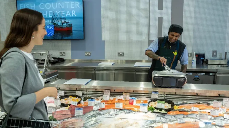 A shopper at a Waitrose fish counter with an employee weighing a seabass