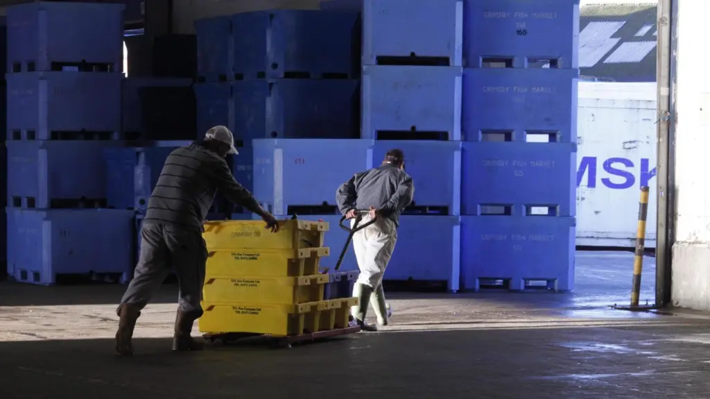 Stacked boxes saying Grimsby Fish Market with two fishermen in the foreground moving smaller yellow crates